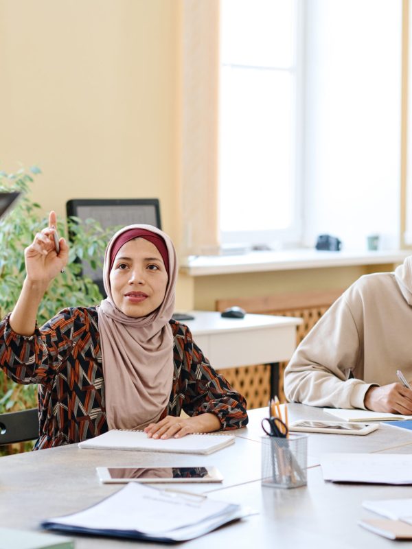 Muslim woman attending language lessons for immigrants raising hand to ask teacher question about task